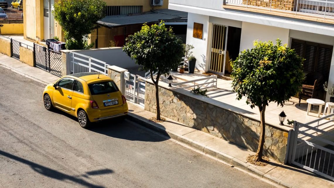 Cozy urban street scene featuring a parked yellow car under bright sunlight, beside modern houses.