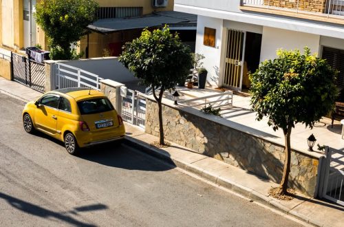 Cozy urban street scene featuring a parked yellow car under bright sunlight, beside modern houses.